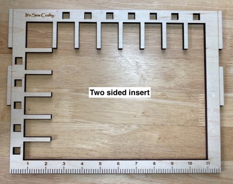 Puede incluir: Una herramienta de costura de madera con un marco rectangular y varias formas recortadas. La herramienta tiene una regla a lo largo del borde inferior, marcada de 1 a 11 pulgadas (aproximadamente 28 cm). El texto "It's Sew Crafty" y "Two sided insert" son visibles.