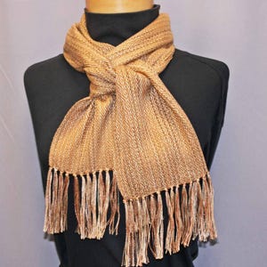 Puede incluir: Bufanda tejida en tonos dorados y marrones, exhibida en un maniqu&iacute;. La bufanda presenta un patr&oacute;n de diamantes y flecos en los extremos. La bufanda est&aacute; anudada en el cuello, mostrando su textura y dise&ntilde;o.