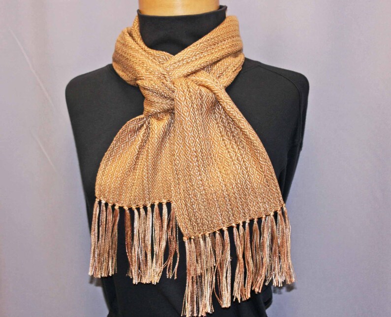 Puede incluir: Bufanda tejida en tonos dorados y marrones, exhibida en un maniqu&iacute;. La bufanda presenta un patr&oacute;n de diamantes y flecos en los extremos. La bufanda est&aacute; anudada en el cuello, mostrando su textura y dise&ntilde;o.