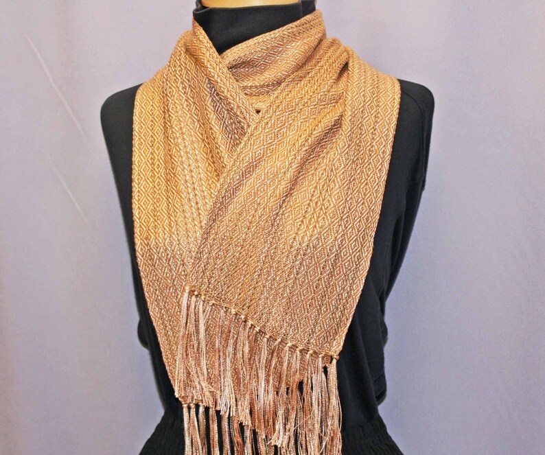 Puede incluir: Bufanda tejida en tonos dorados y beige, con un dise&ntilde;o de rombos. La bufanda tiene un borde con flecos y est&aacute; drapeada sobre un maniqu&iacute; negro. Este accesorio es adecuado para a&ntilde;adir calidez y estilo.