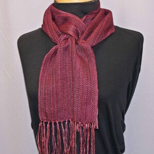 Puede incluir: Bufanda burdeos con estampado de espiga y flecos. La bufanda est&aacute; drapeada sobre un cuello alto negro. El color de la bufanda es una mezcla de tonos rojos y morados, creando una apariencia texturizada. La franja cuelga en la parte inferior de la bufanda.