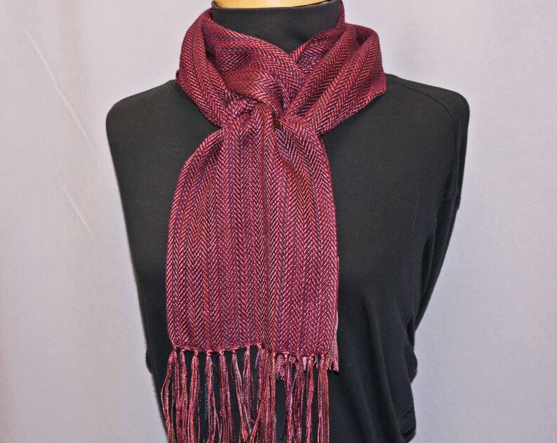 Puede incluir: Bufanda burdeos con estampado de espiga y flecos. La bufanda est&aacute; drapeada sobre un cuello alto negro. El color de la bufanda es una mezcla de tonos rojos y morados, creando una apariencia texturizada. La franja cuelga en la parte inferior de la bufanda.