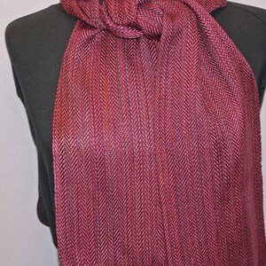 Puede incluir: Bufanda burdeos con estampado de espiga. La bufanda est&aacute; drapeada sobre un maniqu&iacute; negro, mostrando su textura y variaciones de color. La tela parece ser tejida, con tonos de rojo y morado.