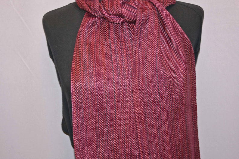 Puede incluir: Bufanda burdeos con estampado de espiga. La bufanda est&aacute; drapeada sobre un maniqu&iacute; negro, mostrando su textura y variaciones de color. La tela parece ser tejida, con tonos de rojo y morado.