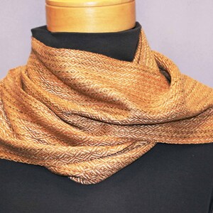 Puede incluir: Bufanda tejida en tonos marrones y dorados, drapeada alrededor de una forma negra. La bufanda presenta un patr&oacute;n de diamantes y una apariencia texturizada, lo que sugiere un accesorio c&aacute;lido y acogedor. La bufanda mide unos 152 cm de largo.