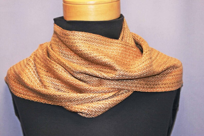 Puede incluir: Bufanda tejida en tonos marrones y dorados, drapeada alrededor de una forma negra. La bufanda presenta un patr&oacute;n de diamantes y una apariencia texturizada, lo que sugiere un accesorio c&aacute;lido y acogedor. La bufanda mide unos 152 cm de largo.