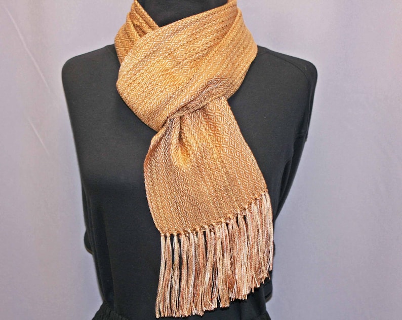 Puede incluir: Bufanda tejida en tonos dorados y marrones, exhibida en un maniqu&iacute; negro. La bufanda presenta un patr&oacute;n de diamantes y flecos en los extremos. La bufanda est&aacute; hecha de un material suave y texturizado. Mide unos 170 cm.