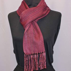 Puede incluir: Una bufanda burdeos con un patr&oacute;n de espiga y flecos. La bufanda est&aacute; drapeada sobre un maniqu&iacute; negro. El color de la bufanda es una mezcla de tonos rojos y morados, con un aspecto texturizado. Los flecos en la parte inferior a&ntilde;aden un elemento decorativo.