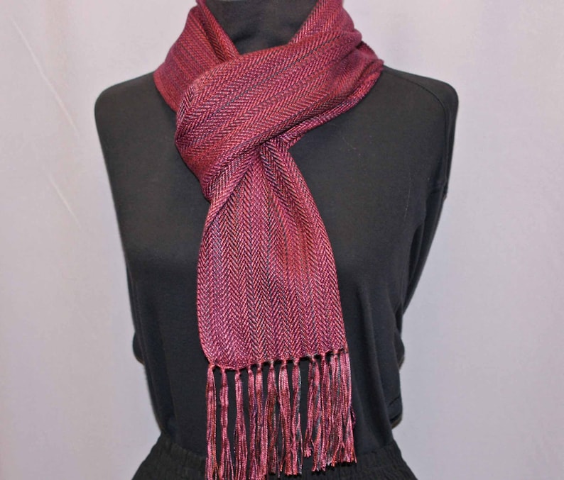 Puede incluir: Una bufanda burdeos con un patr&oacute;n de espiga y flecos. La bufanda est&aacute; drapeada sobre un maniqu&iacute; negro. El color de la bufanda es una mezcla de tonos rojos y morados, con un aspecto texturizado. Los flecos en la parte inferior a&ntilde;aden un elemento decorativo.