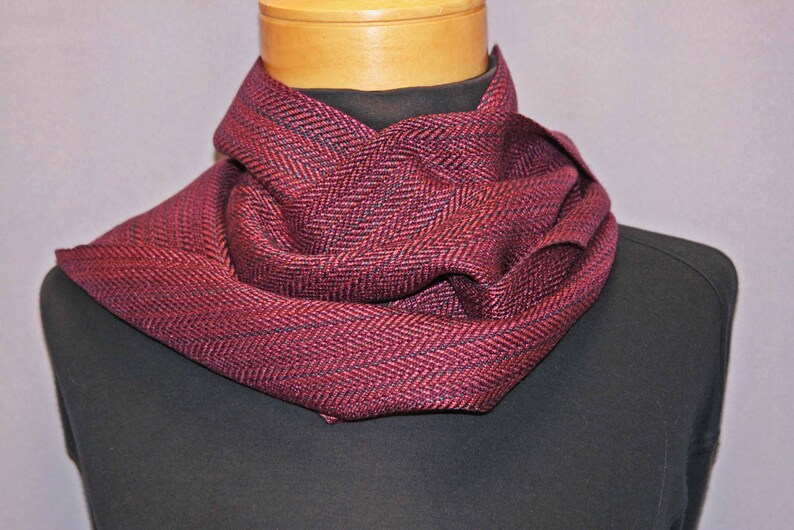 Puede incluir: Una bufanda burdeos y negra con estampado de espiga est&aacute; drapeada alrededor del cuello de un maniqu&iacute;. La bufanda est&aacute; hecha de un material suave y tejido y tiene un aspecto texturizado. Un accesorio elegante para el clima fr&iacute;o.