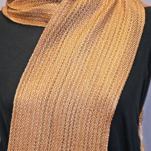 Puede incluir: Bufanda tejida en tonos dorados y marrones, sobre una prenda negra. La bufanda presenta un patr&oacute;n de diamantes repetido, creando una apariencia texturizada. El degradado de color cambia de claro a oscuro.