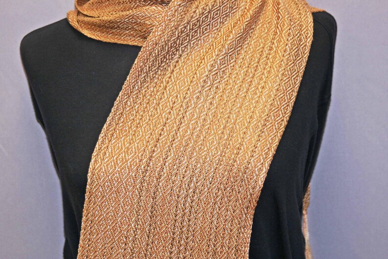 Puede incluir: Bufanda tejida en tonos dorados y marrones, sobre una prenda negra. La bufanda presenta un patr&oacute;n de diamantes repetido, creando una apariencia texturizada. El degradado de color cambia de claro a oscuro.