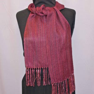 Puede incluir: Bufanda burdeos con estampado de espiga y flecos. La bufanda est&aacute; drapeada sobre un maniqu&iacute; negro. La bufanda est&aacute; hecha de una tela tejida y tiene un aspecto texturizado. La franja est&aacute; en la parte inferior de la bufanda.