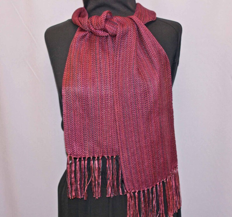 Puede incluir: Bufanda burdeos con estampado de espiga y flecos. La bufanda est&aacute; drapeada sobre un maniqu&iacute; negro. La bufanda est&aacute; hecha de una tela tejida y tiene un aspecto texturizado. La franja est&aacute; en la parte inferior de la bufanda.