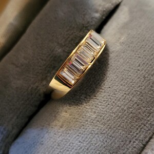 May include: A gold band ring featuring a channel setting with five clear baguette-cut stones arranged in a row. The metal has a polished finish, and the stones are rectangular in shape, reflecting light across their faceted surfaces.