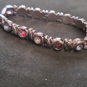 May include: A silver-toned bracelet with an alternating pattern of XOXO links and round, colorful gemstones. The gemstones are pink, clear, red, and light blue. The bracelet is on a dark green surface.