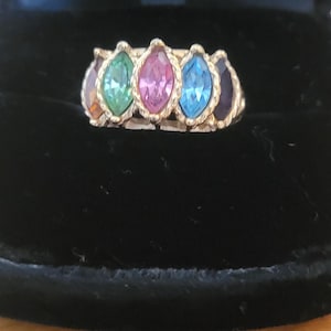 May include: A gold-toned ring featuring five marquise-cut gemstones arranged in a row. The stones display a vibrant spectrum of colors, including orange, green, pink, light blue, and deep purple, all set within a textured, rope-style metal band.