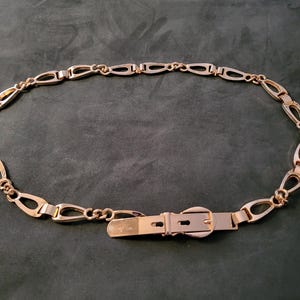 May include: Gold-toned metal belt with an oval link chain design. The belt features a buckle closure and a series of elongated oval links connected by smaller chain segments. The belt is displayed on a dark green surface.