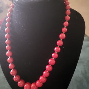 May include: A red beaded necklace displayed on a black mannequin. The necklace features large, round beads in a vibrant red color, strung together with silver-toned spacers. The beads graduate in size, creating a visually appealing design.