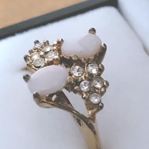 May include: A gold-toned ring featuring a cluster design. The ring has two oval, white stones and several smaller clear stones. The ring is set against a white background.