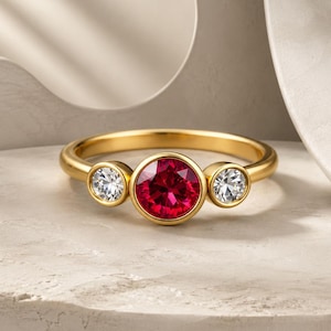 Könnte beinhalten: Ein goldener Ring mit einem großen, runden, roten Edelstein in der Mitte, flankiert von zwei kleineren, klaren Edelsteinen. Der Ring hat ein schlichtes, elegantes Design, perfekt für den Alltag oder besondere Anlässe.