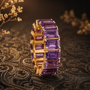 Op de afbeelding: Een eeuwigheidsring met een doorlopende rij rechthoekige, smaragdgeslepen paarse amethist edelstenen in een geelgouden metalen zetting. De ring toont een herhalend patroon van levendige violette stenen die zijn vastgezet met kleine gouden klauwtjes.