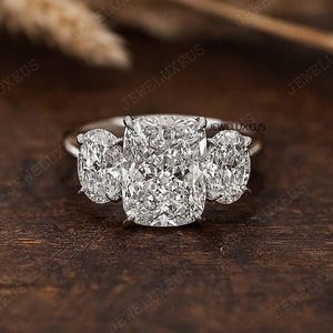 Könnte beinhalten: Ein silberner Verlobungsring mit einem großen, kissenförmigen Mittelstein und zwei oval geschliffenen Seitensteinen. Der Ring liegt auf einer dunklen Holzoberfläche. Die Diamanten funkeln. Ein klassisches Design.