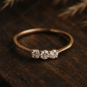 May include: A gold ring with three clear gemstones. The ring has a textured band. The ring is displayed on a dark wooden surface. The gemstones are set in a row. The ring's design is simple and elegant.