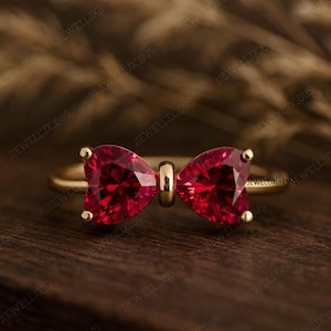 May include: A gold ring featuring a bow-shaped design with two heart-shaped, deep red gemstones. The ring is set on a wooden surface, showcasing the jewelry's intricate details and vibrant color.