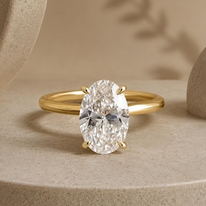 May include: A gold ring with a large, oval-cut diamond. The diamond is the focal point, set with four prongs on a simple, polished gold band. The ring is displayed on a neutral-toned surface, with soft lighting.