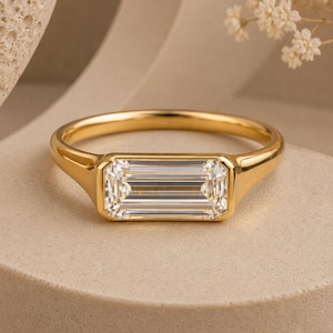Op de afbeelding: Een gouden ring met een rechthoekige, heldere edelsteen. De ring heeft een eenvoudige band en een bezel setting die de edelsteen vasthoudt. De ring wordt gepresenteerd op een neutraal getinte ondergrond.