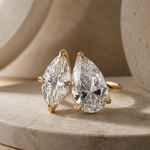 Könnte beinhalten: Ein goldener Ring mit zwei tropfenförmigen Diamanten. Die Diamanten sind nebeneinander angeordnet und im Brillantschliff geschliffen. Der Ring wird auf einer neutralen Oberfläche präsentiert.