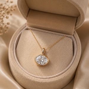 May include: A gold pendant necklace with an oval-shaped, clear gemstone set in a gold bezel. The necklace is displayed in an open, beige jewelry box. The box sits on a satin-like, beige surface. The necklace is a piece of fine jewelry.