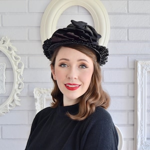 May include: A woman wearing a black mock neck shirt and a black hat with a large, ruffled brim. The hat is decorated with black satin ribbons and lace.