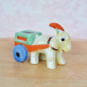 May include: A ceramic goat pulling a small cart. The goat is primarily yellow with brown accents and orange horns. The cart is green and orange with a blue wheel. The figurine has a glossy finish.