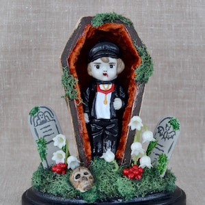 May include: A small porcelain doll dressed as a vampire is lying in a wooden coffin. The coffin is surrounded by moss, white flowers, and red berries. There are two gray tombstones with inscriptions on either side of the coffin.