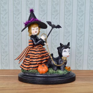May include: A Halloween-themed figurine featuring a witch in a black and orange striped dress and hat, holding a bat-shaped wand and a leash attached to a black lamb. A small orange pumpkin and a bowl of candy are also present.