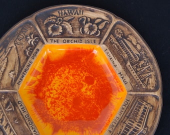 May include: A hexagonal ceramic ashtray with a yellow and orange glaze. The ashtray is divided into six sections, each with a different Hawaiian landmark and its name. The landmarks include Diamond Head, Pearl Harbor, Tolanai Palace, Lahaina Maui, Menehune Pond, and Kauai. The text "The Orchid Isle" is printed in the center of the ashtray.