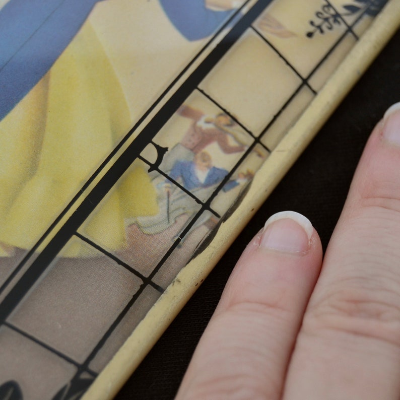 K&ouml;nnte beinhalten: Ein Vintage-Blechtablett mit einem schwarz-wei&szlig;en Gittermuster und einem verblassten Bild einer Frau in einem gelben Kleid und einem blauen Kleid. Der Text "Best" ist in der oberen rechten Ecke des Tabletts sichtbar.