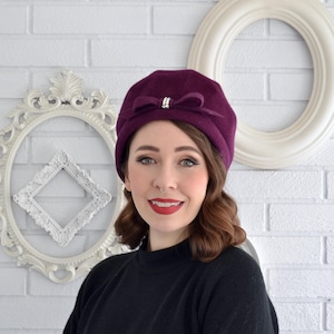 May include: A deep purple cloche hat with a bow and rhinestone accent. The hat is made of a soft material and has a classic, vintage style. The hat is a stylish accessory for any outfit.