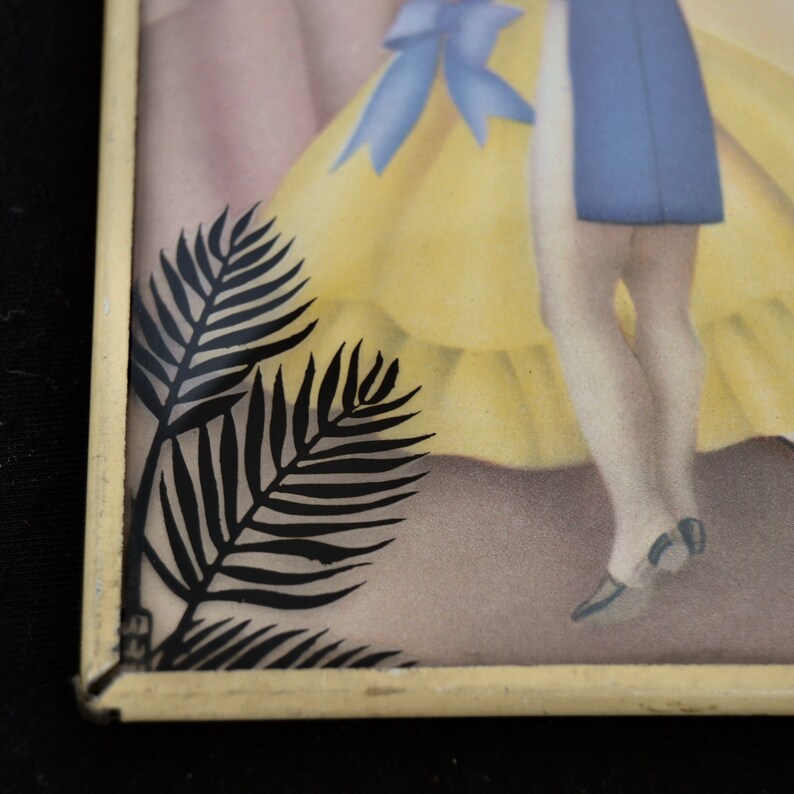 K&ouml;nnte beinhalten: Ein gerahmtes Bild einer Frau in einem gelben Kleid und einer blauen Sch&auml;rpe, mit einer schwarzen Silhouette einer Palmenwedel im Vordergrund.