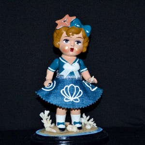 May include: A vintage doll figurine in a sailor outfit with a denim skirt, holding a starfish. The doll has a blue bow and a starfish hair accessory. The base features coral and a beach scene.