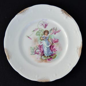 May include: A white porcelain plate with a gold rim features a hand-painted design of a fairy in a blue dress surrounded by pink and purple flowers.