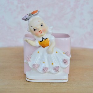 May include: A vintage ceramic figurine of a girl wearing a white dress with pink flowers and a yellow bodice. She is holding a pink flower in her hand and has a pink flower on her head. The figurine is a planter with a pink square base.