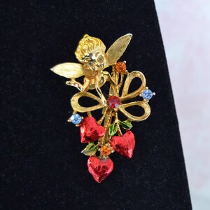 May include: A gold-coloured brooch depicting a cherub with blue eyes and wings, holding a bouquet of red heart-shaped charms. The brooch is decorated with red, blue, and orange gemstones, and green leaves. The brooch is set against a black background.