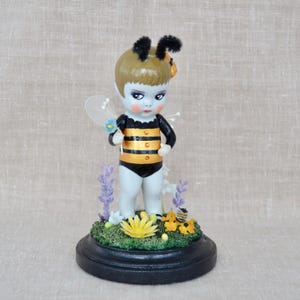 May include: A ceramic figurine of a child dressed as a bee, with black and gold striped bodysuit, wings, and antennae. The figurine holds a blue flower and stands on a base with flowers and a small bee. The base is black.