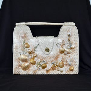 May include: A structured white handbag featuring a textured surface adorned with various seashells, gold glitter accents, and small white floral embellishments. The bag includes a top handle and a central flap closure with a gold-toned metal turn lock.