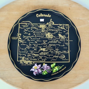May include: A round black metal tray featuring a gold-coloured map of Colorado with various city names, landmarks, and illustrations. The word Colorado is printed at the top, and a colourful illustration of the Colorado Columbine flower is at the bottom.