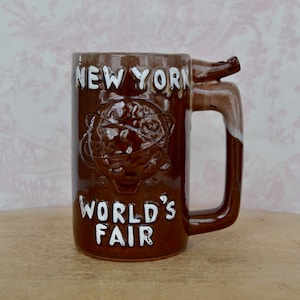 May include: A brown ceramic mug with a white handle and a white inscription that reads "New York World's Fair". The mug has a brown and white glaze and a 3D globe design.