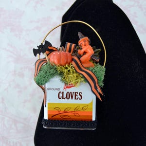 May include: A decorative ornament featuring a vintage white tin labeled GROUND CLOVES with orange and brown stripes. It is topped with green moss, an orange pumpkin, a black bat cutout, a small orange witch figure, and orange and black ribbon.
